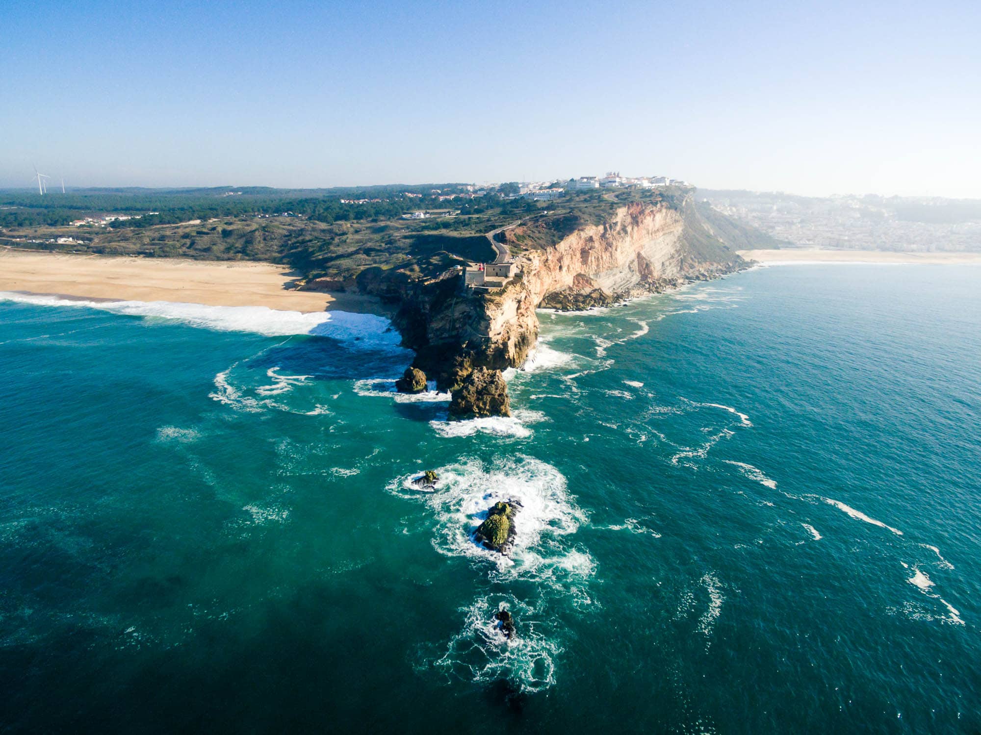 Drone view over the Sítio headland — Praia do Norte sand at bottom-left, rocky headland, wind turbines on the horizon